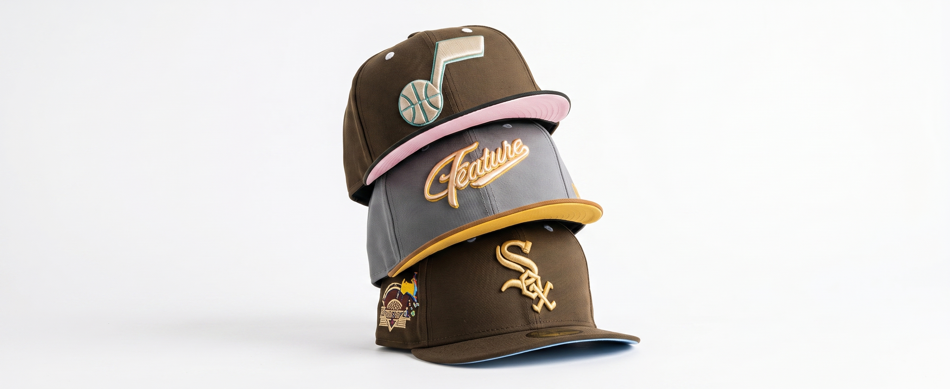A stack of three premium fitted hats featuring a brown Utah Jazz cap, a grey Feature script cap, and a brown Chicago White Sox cap with a side patch.