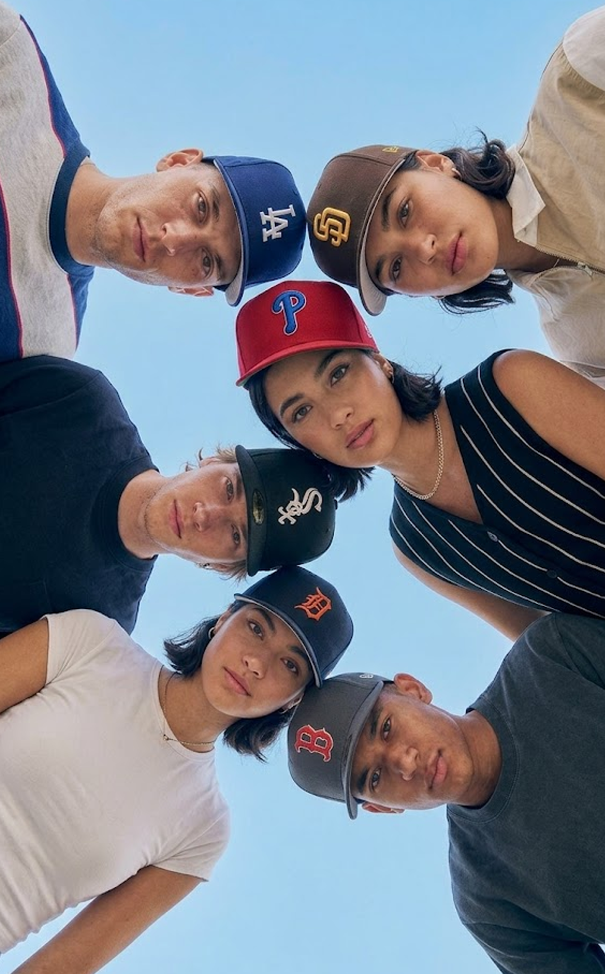 Diverse group of young adults looking down at the camera wearing various New Era MLB baseball caps against a clear blue sky.