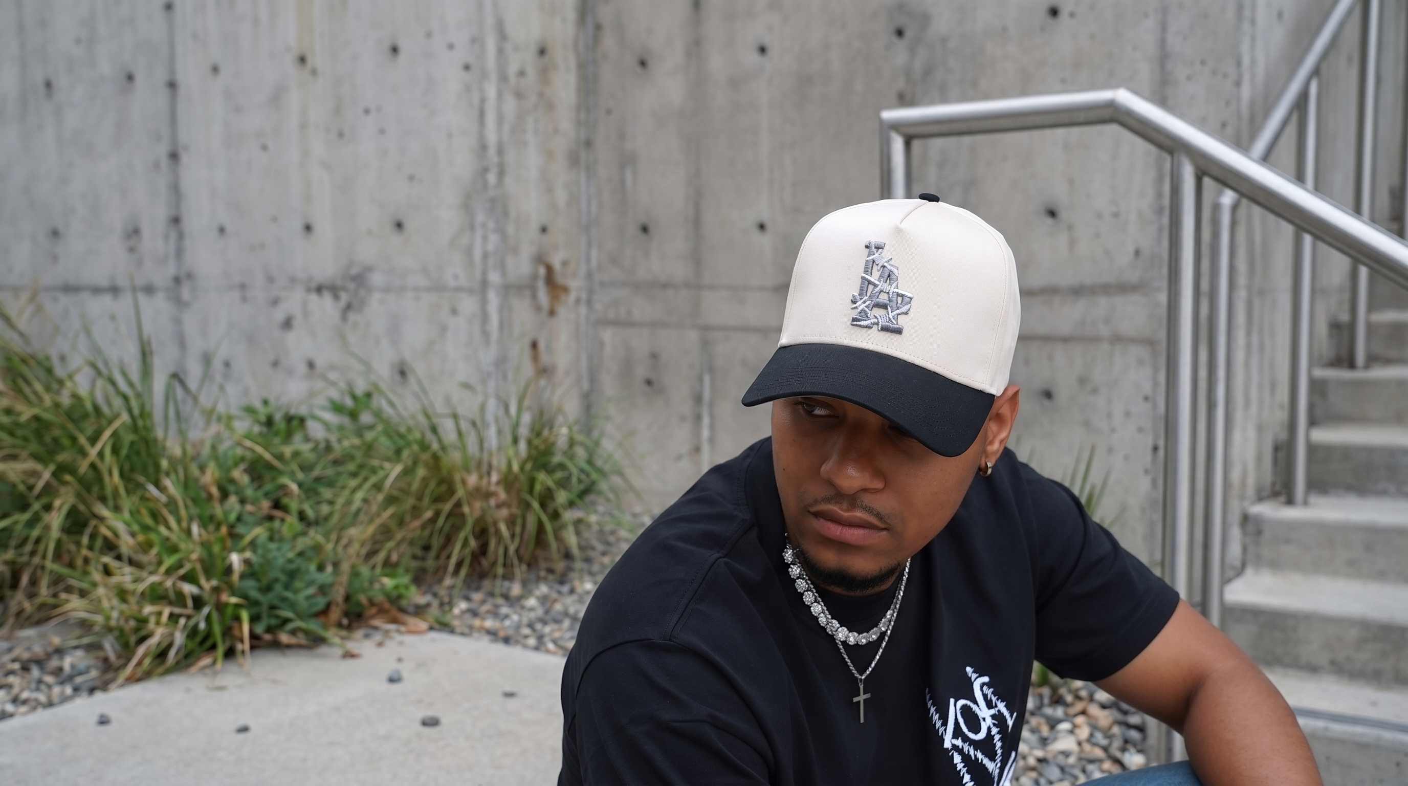 Man wearing a custom cream and black Los Angeles cap with a stylized metallic logo, sitting outdoors on concrete stairs