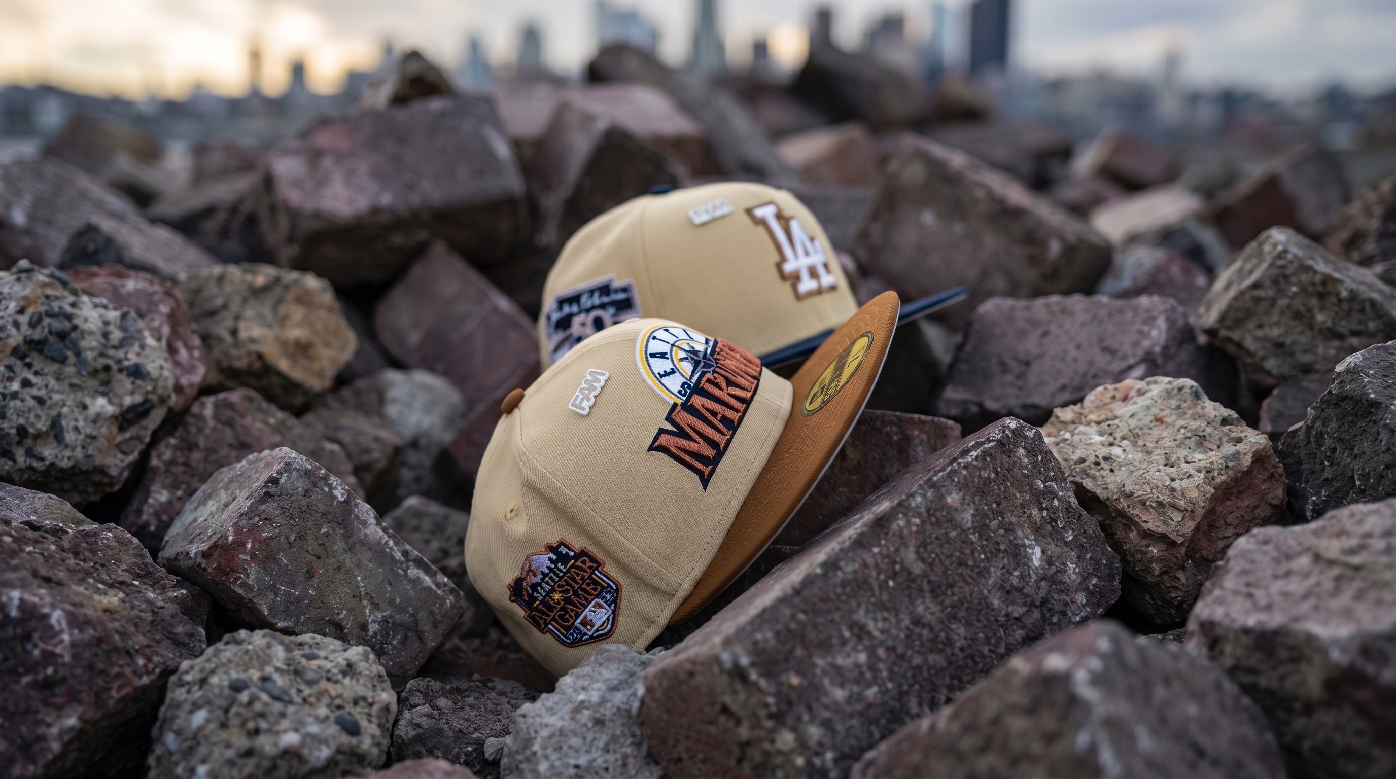 Custom beige and brown Seattle Mariners and Los Angeles Dodgers fitted hats with side patches, resting on rocks against a blurred city skyline