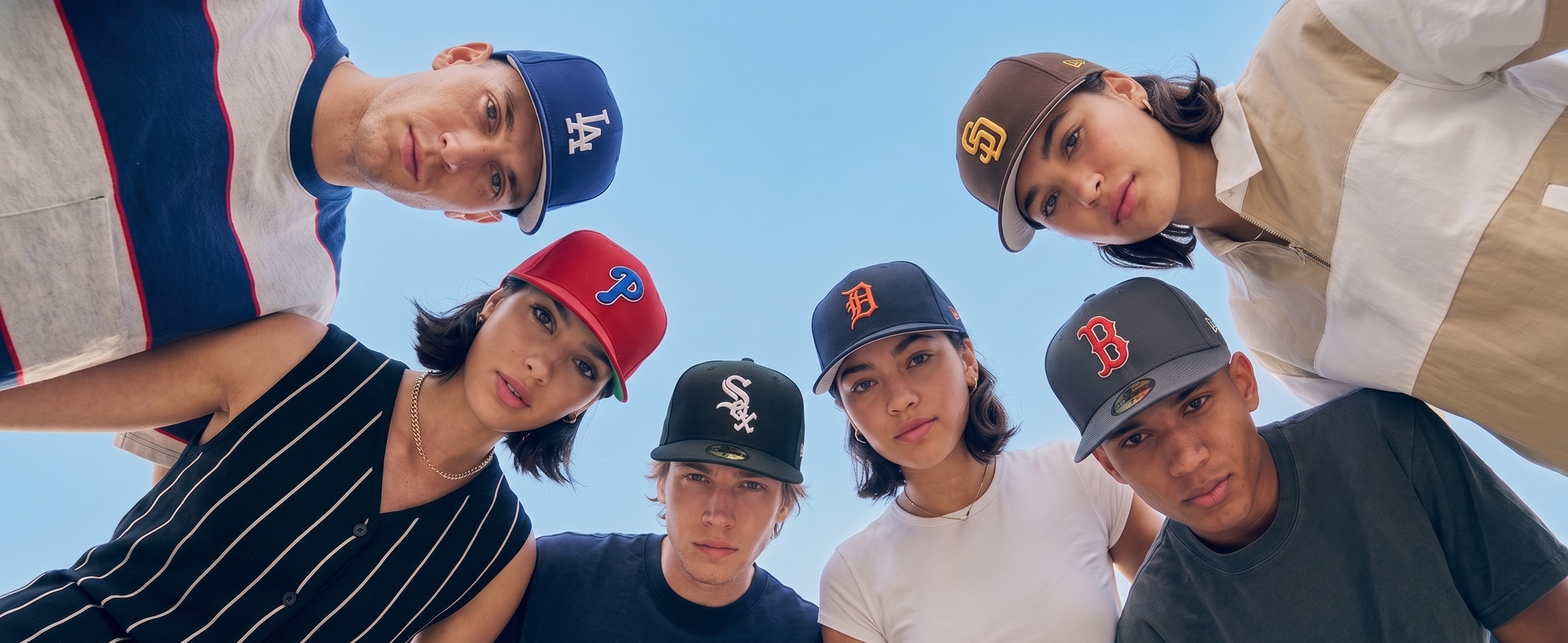 Diverse group of young adults looking down at the camera wearing various New Era MLB baseball caps against a clear blue sky.