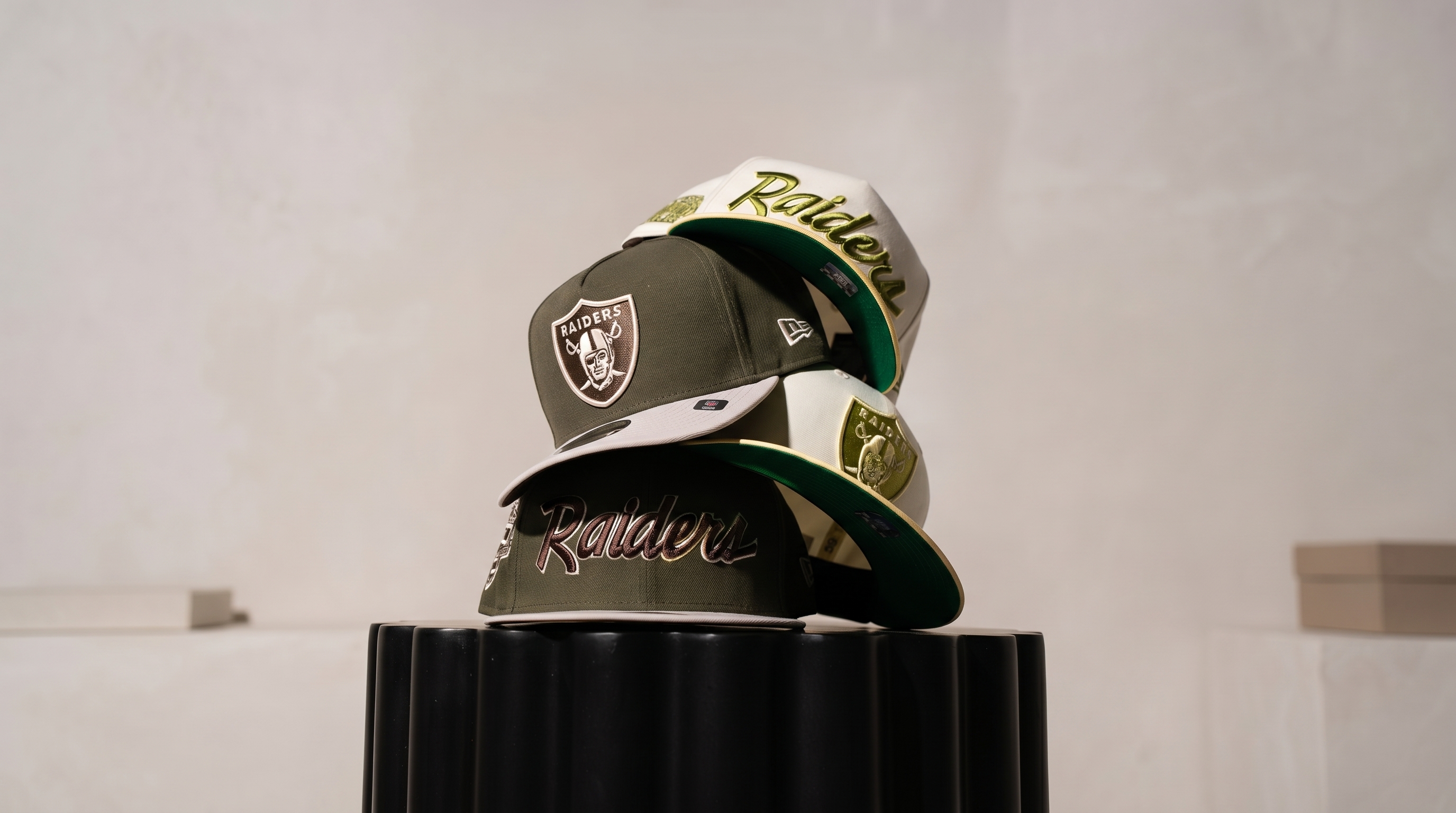 Stack of three custom Raiders hats in olive green and cream colorways, displayed on a black pedestal against a minimalist beige wall